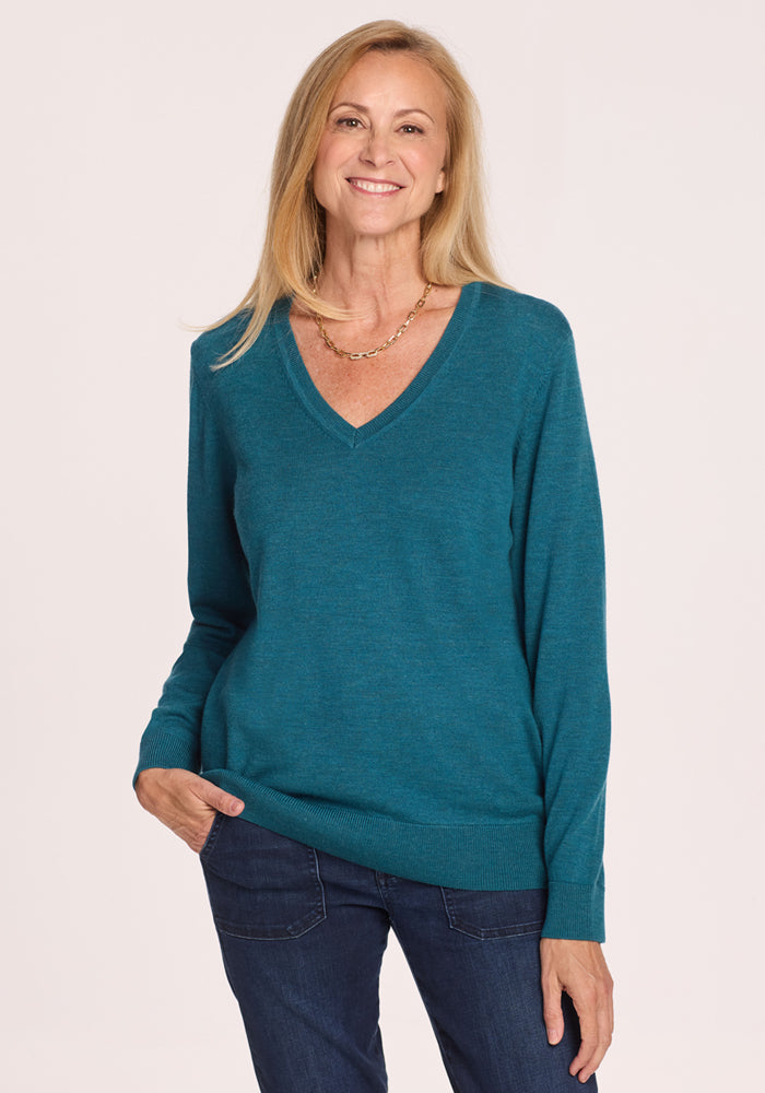 A smiling woman with straight blonde hair wears the Woolx Francesca Sweater in Harbor Teal, paired with dark jeans and a gold chain necklace. She stands against a plain light background with one hand in her pocket. 