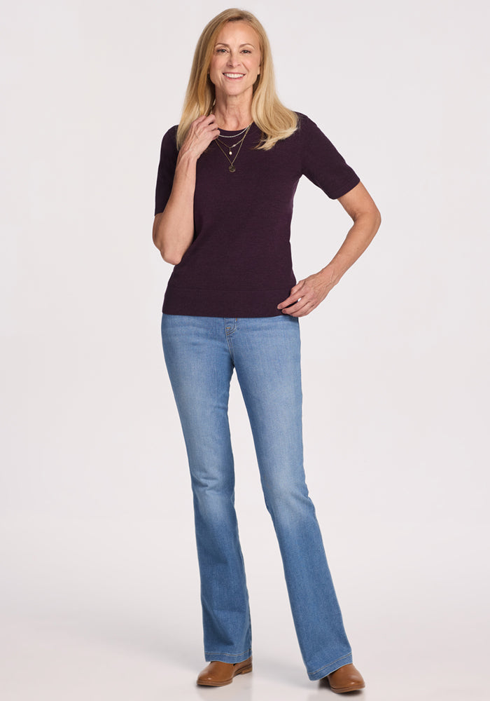 A smiling woman with long blonde hair stands against a white background, wearing the Woolx Anna Crew Sweater in Deep Plum, paired with blue jeans, brown shoes, and layered necklaces. One hand rests on her hip; the other touches her necklace.