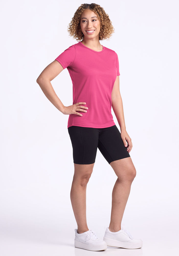 A person with curly hair smiles in a relaxed pose, wearing the Woolx Liza Crew Neck Tee in Raspberry Fizz, black bicycle shorts, and white sneakers against a plain white background.