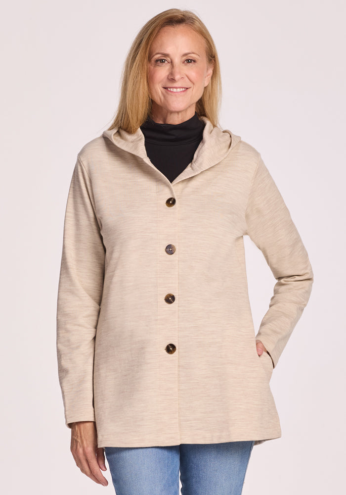 A smiling woman with long blonde hair wears the Woolx Edna Hooded Cardigan in Cream Heather over a black turtleneck and blue jeans, standing against a light background. This cozy piece is perfect for cold weather. 