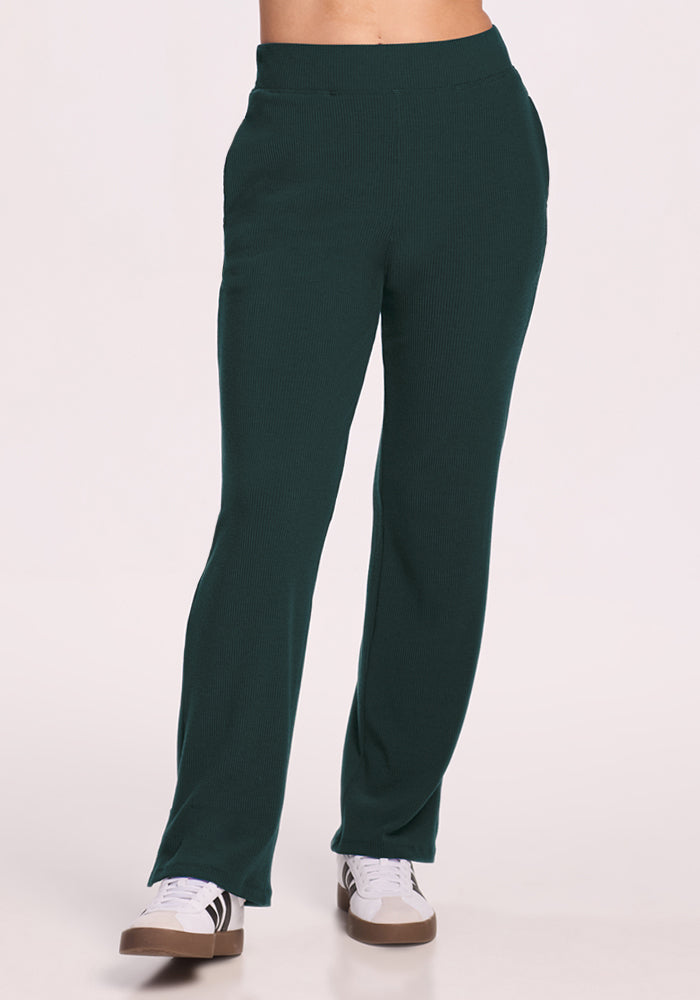 A person wears Woolx Luca Ribbed Pants in Deep Timber and white sneakers with black stripes, standing against a plain light background. The upper body is partially out of frame. 