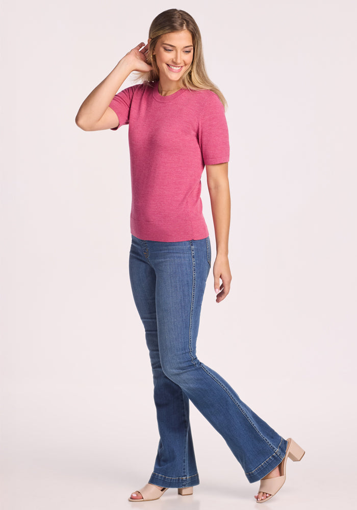 A woman with long light brown hair smiles, wearing the Woolx Anna Crew Sweater in Wild Salmon, blue bootcut jeans, and beige heeled sandals. She holds one hand near her ear against a plain, light background.