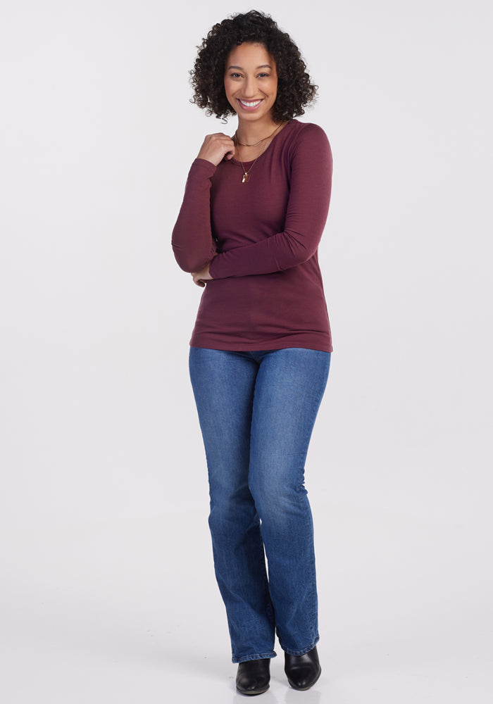 A curly-haired woman smiles against a white background, wearing the Woolx Remi Long Sleeve T Shirt in Grape Harvest, blue jeans, black boots, a necklace, and touching her chin with one hand.