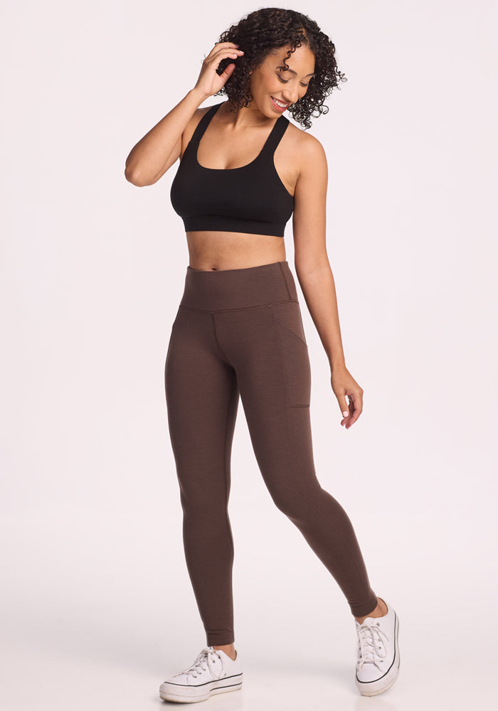 A woman with curly hair wears a black sports bra and Woolx Piper Pocket Petite leggings in Bracken while smiling and looking down, standing against a plain white background.