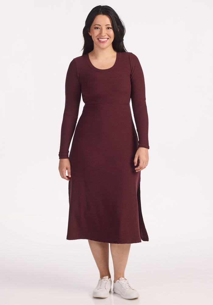 A woman with long dark hair, wearing the Woolx Caroline Ribbed Twirl Dress in Toasted Garnet and white sneakers, smiles against a plain white background. 