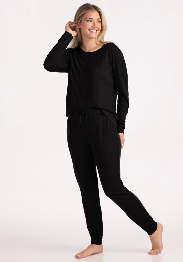 A woman stands barefoot against a light background, smiling and touching her hair while wearing Woolx Mary Kate Lounge Pants in black with a matching long-sleeve top—perfect for effortless travel and loungewear.