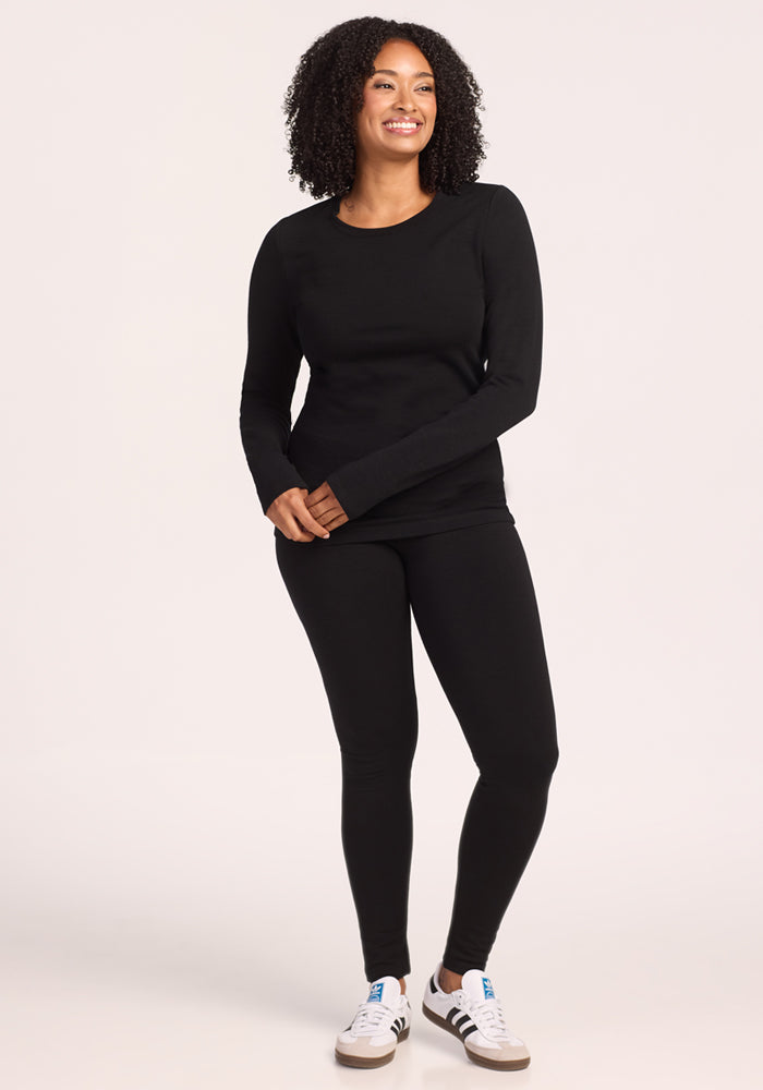 A woman with curly hair smiles while wearing the Woolx Sadie Baselayer Top in black, paired with matching leggings and white sneakers with black stripes, standing against a plain light background.
