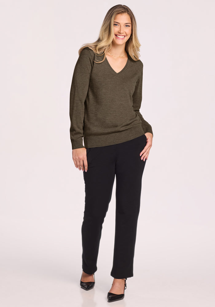 A woman with long blonde hair smiles against a light background, wearing the Woolx Francesca Sweater in Pine Needle, paired with black pants and heels. One hand is in her pocket while the other rests by her side.