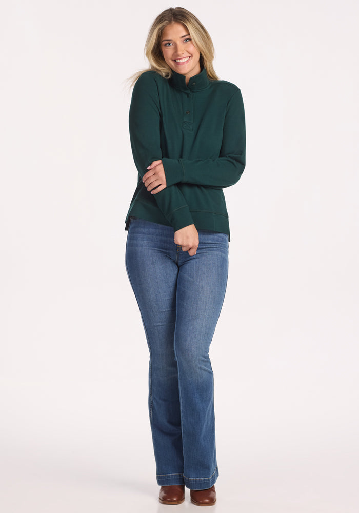A woman in the Woolx Annie 3 Button Pullover in Deep Timber and blue jeans stands smiling with arms crossed against a plain white background. She has light brown hair and is wearing brown shoes.