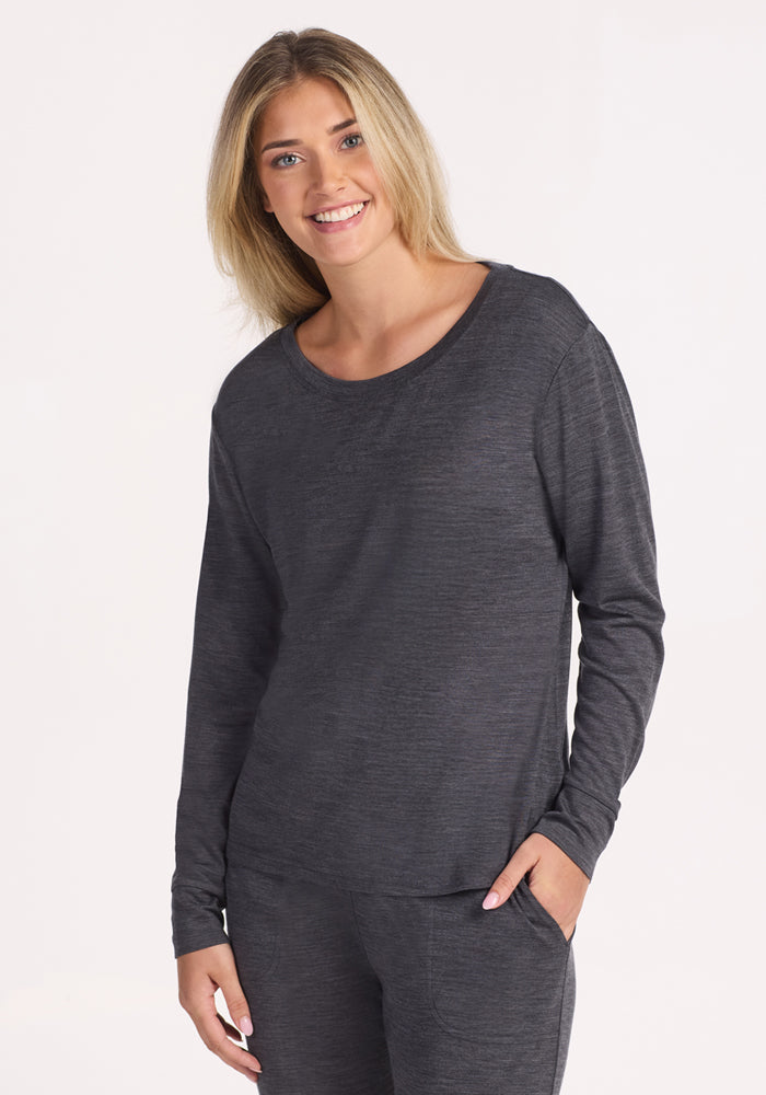 A person with long blonde hair wears the Woolx Ashlie Lounge Top in Cinder Melange, smiling with one hand in their pocket against a plain white background. 