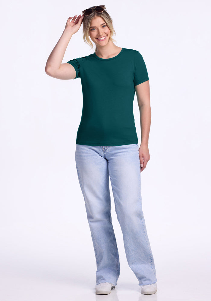 A smiling woman stands against a plain background, wearing the Woolx Liza Crew Neck Tee in Riverbank Green, paired with light blue jeans and white shoes. She holds sunglasses on her head with one hand.