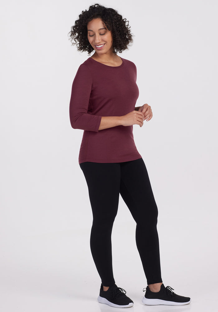 A woman with curly hair smiles while looking downward, wearing the Woolx Jenny 3/4 Sleeve Crew Neck in Grape Harvest, black leggings, and black sneakers against a plain white background.