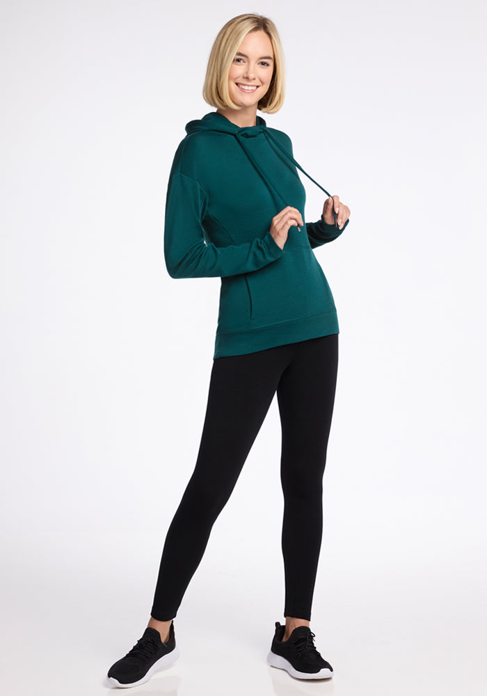 A blonde woman smiles while posing in the Woolx Callie Hoodie in Riverbank Green, made from ultra-soft fabric. She pairs it with black leggings and sneakers, holding the hoodie drawstrings with both hands against a plain white background.
