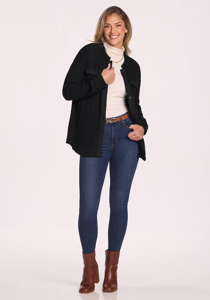 A woman smiles while standing against a white background, wearing the Woolx Oaklynn Waffle Shacket in black over a cream turtleneck, blue skinny jeans, brown belt, and brown ankle boots.