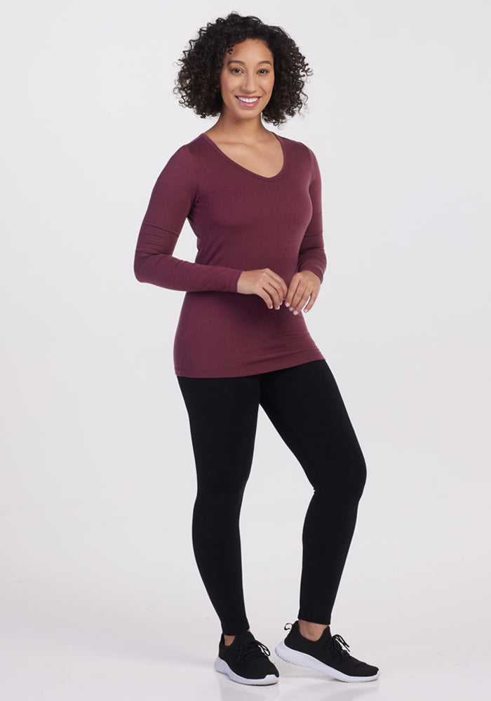 A woman with curly hair smiles while standing, wearing the Woolx Layla V Neck Top in Grape Harvest, black leggings, and black sneakers with white soles against a plain white background.