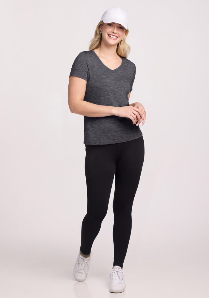 A woman smiles while walking, wearing the Woolx Junie V Neck in Cinder Melange, a white baseball cap, black leggings, and white sneakers against a plain light background.