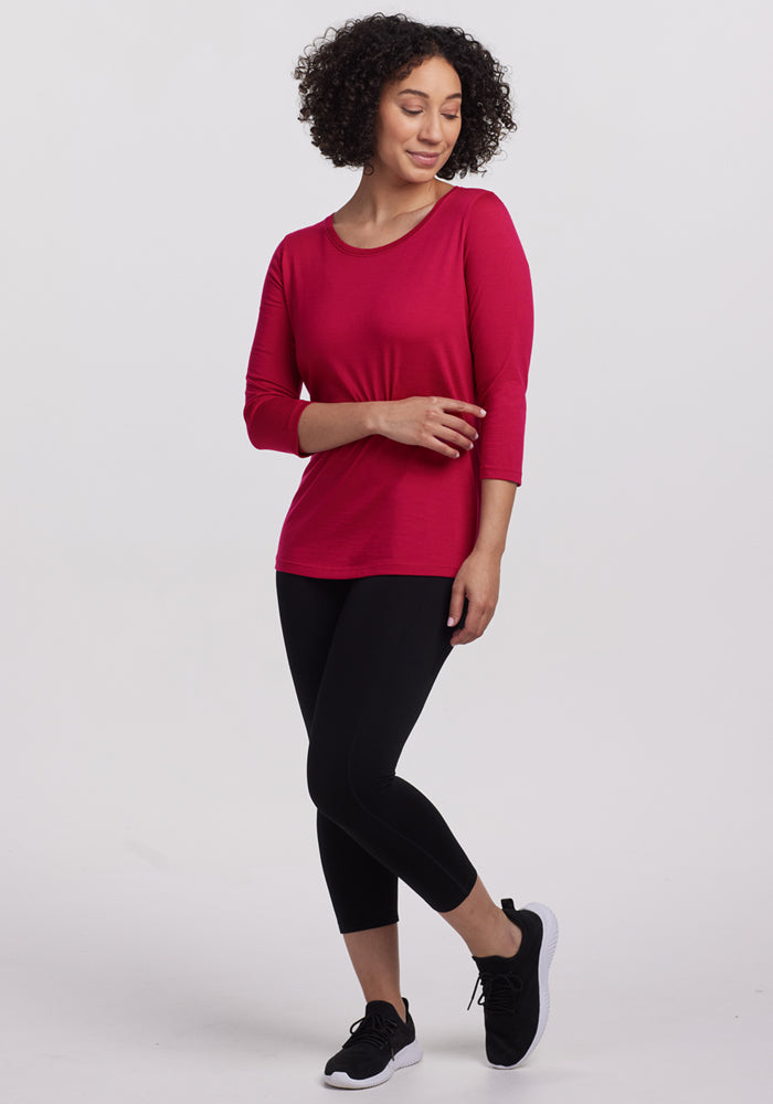 A woman with curly hair wears the Woolx Jenny 3/4 Sleeve Crew Neck in Poppy Spark, paired with black leggings and black athletic shoes. She stands against a white background, looking down with a slight smile and one leg crossed.