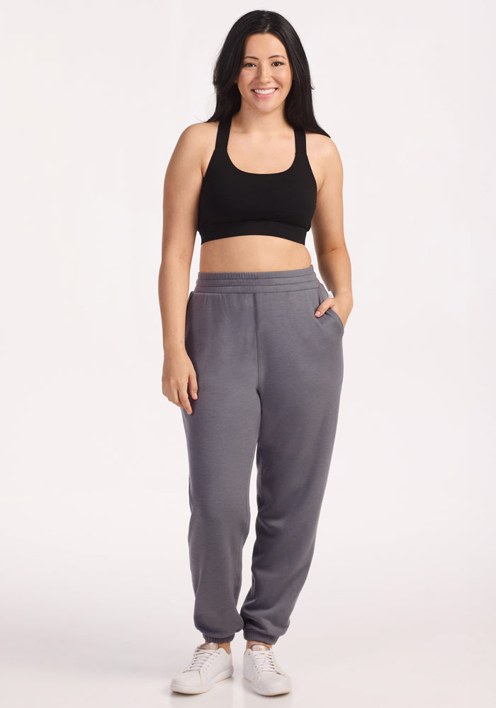 A woman with long dark hair smiles, wearing a black sports bra, Woolx Parker Sweatpants in Arctic Dusk gray, and white sneakers. Her left hand is in her pocket against a plain white background.