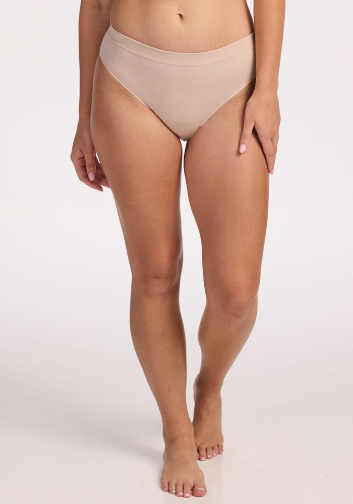 A person stands barefoot on a white background, wearing Woolx Maeve Bikini in Sand. Their body is shown from the waist down, displaying the high-cut, moisture-wicking merino wool underwear with arms relaxed by their sides.