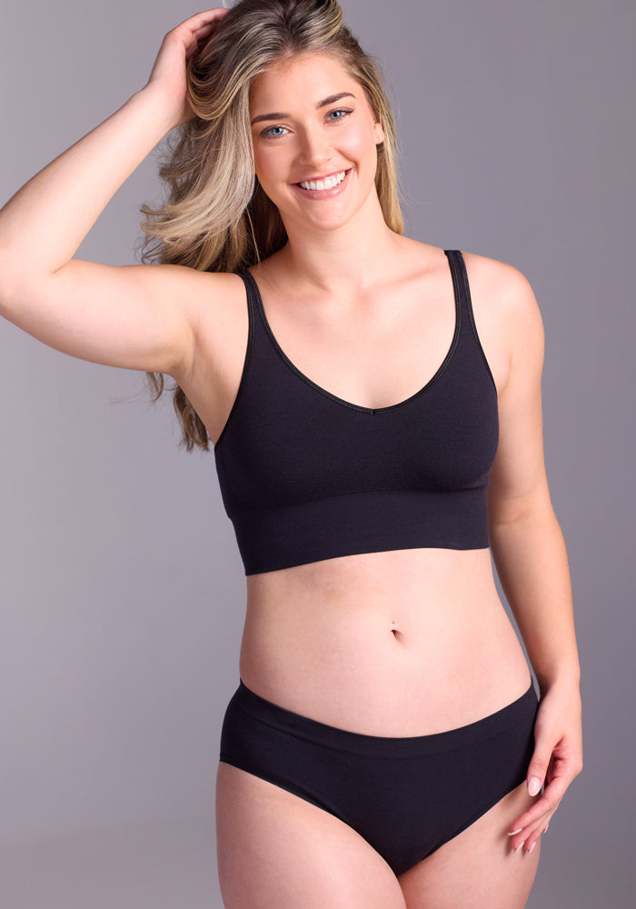 A smiling woman with long, wavy blonde hair poses confidently in Woolx’s Maeve Bikini - Black, paired with a matching black sports bra, against a plain light gray background.