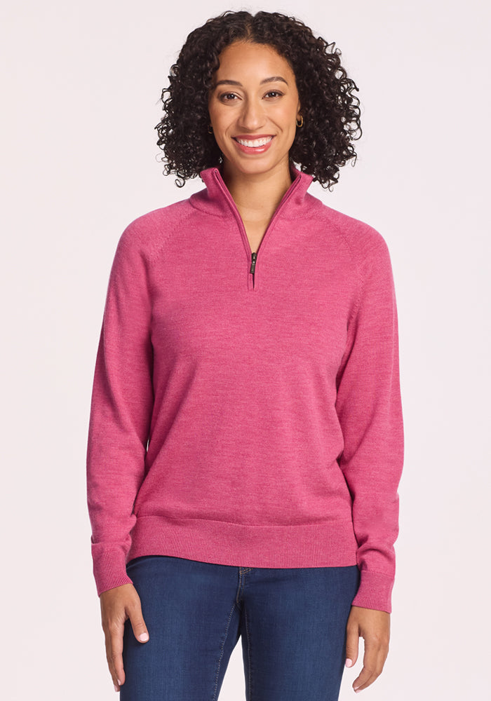 A woman with curly hair is smiling and wearing the Woolx Collins Half Zip Sweater in Wild Salmon and blue jeans while standing against a plain light background. 
