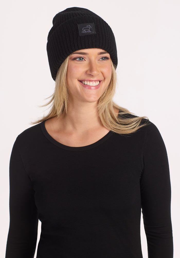 A smiling person models the Woolx Ria Chunky Beanie in black with a long-sleeve shirt against a plain white background.