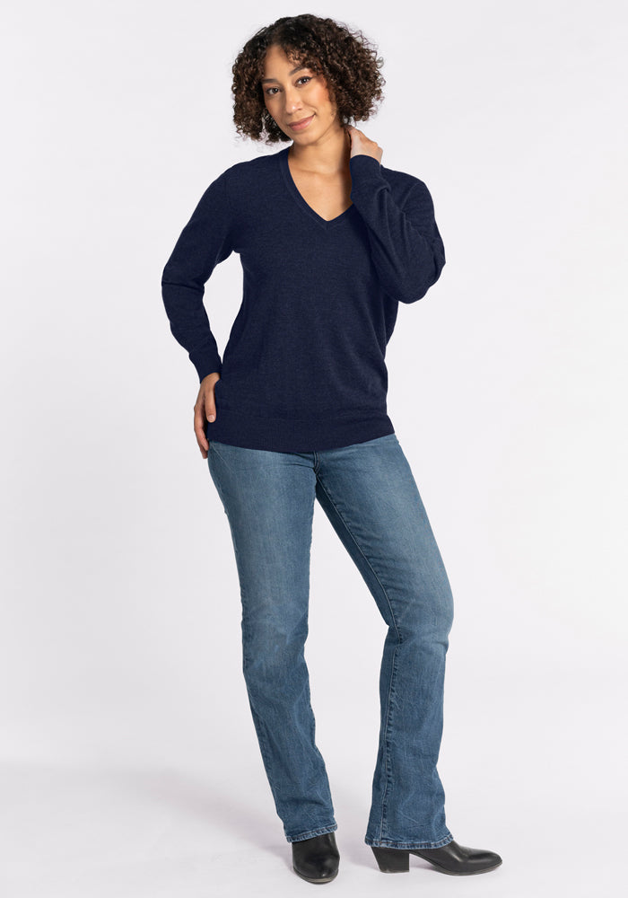 A person with curly hair wears the Woolx Francesca Sweater in Storm Cloud, blue jeans, and black shoes. Against a white background, they pose with one hand on their hip and the other touching their face.