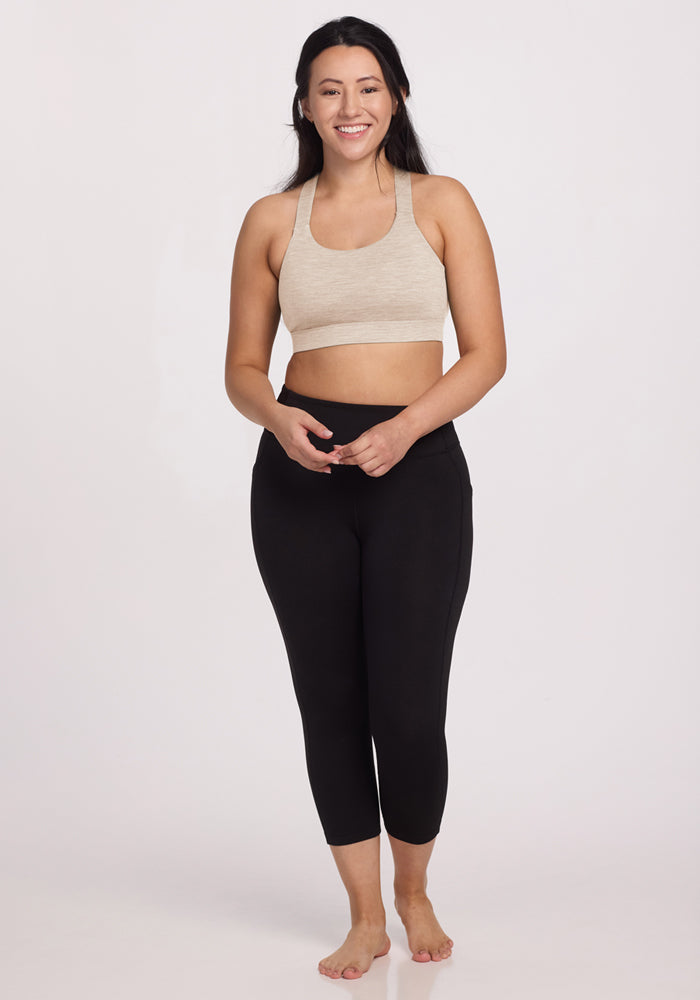 A woman with long dark hair stands barefoot, smiling at the camera. She wears the Woolx Livi Bra in Cream Heather and black high-waisted leggings against a plain light background.