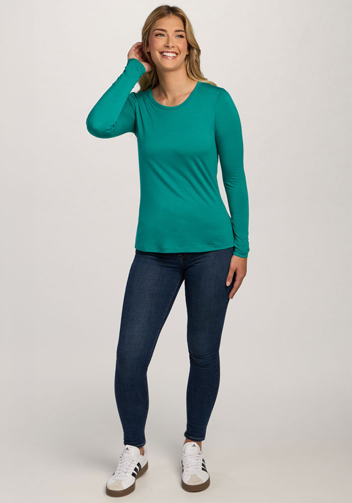 Woman smiles in a Woolx Remi Long Sleeve T-Shirt - Cabana Crush, blue jeans, and white sneakers against a plain background.