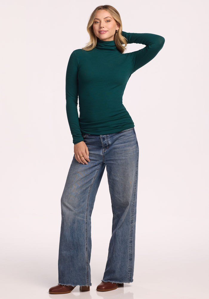 A woman poses against a light background wearing the Woolx Sage Ribbed Turtleneck in Riverbank Green, paired with wide-leg blue jeans and brown shoes. One hand rests on her hip and the other behind her head.
