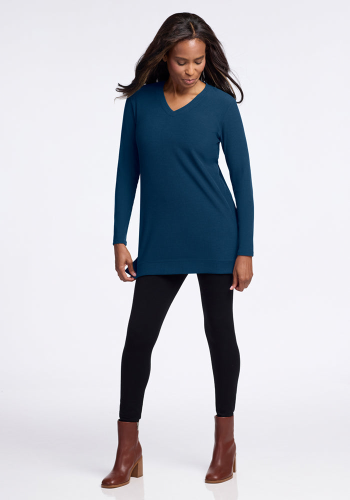 A woman in the Woolx Evie Ribbed Tunic in Majolica Blue, black leggings, and brown ankle boots stands against a white background, looking down and to the side.