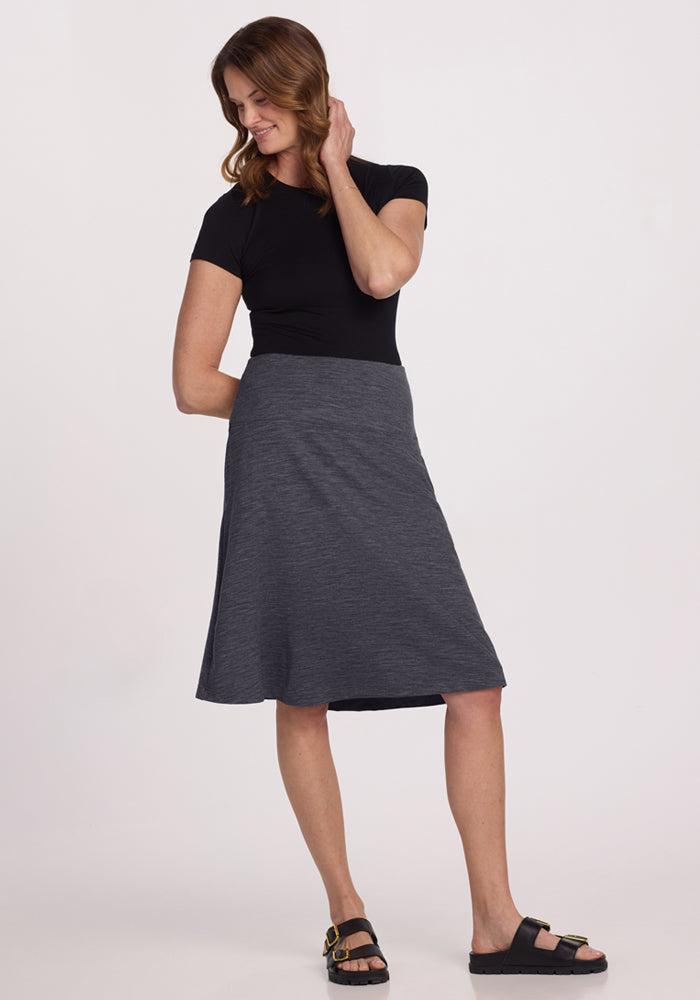 A woman stands against a plain background wearing a black short-sleeve top, black sandals, and the Woolx Marilyn Flowy Midi Skirt in Cinder Melange. She is smiling, looking down, with one hand touching her hair.