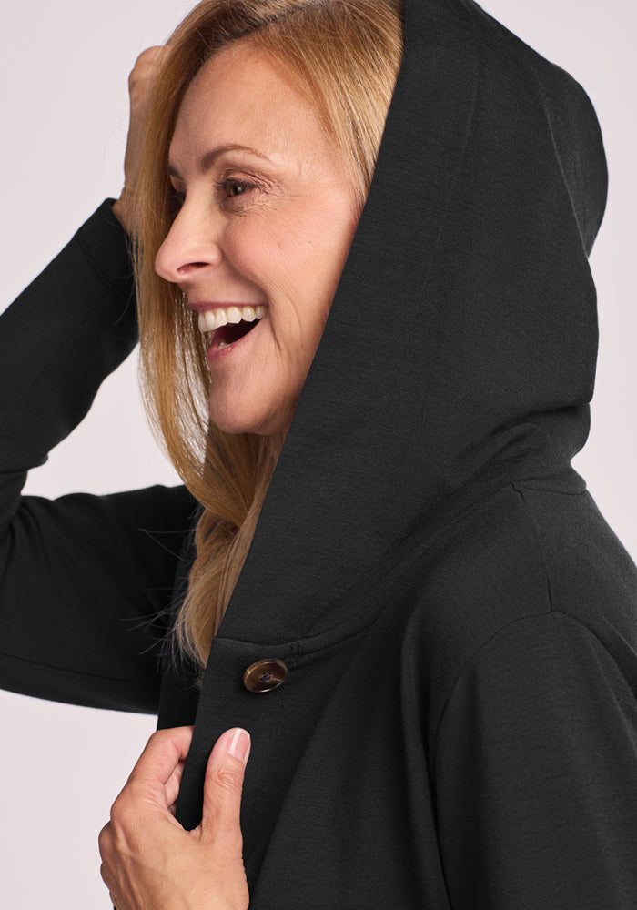 A smiling blonde woman wears the Woolx Edna Hooded Cardigan in black, featuring a button detail, shown in a close-up side profile—ideal as a cozy cold weather sweater.