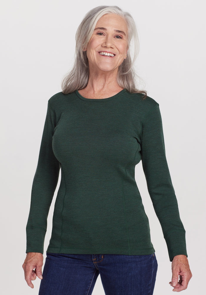 An older woman with long gray hair smiles while standing against a white background. She wears the Woolx Hannah Long Sleeve Top in Forest, a dark green, moisture-wicking Merino wool base layer, paired with blue jeans. 