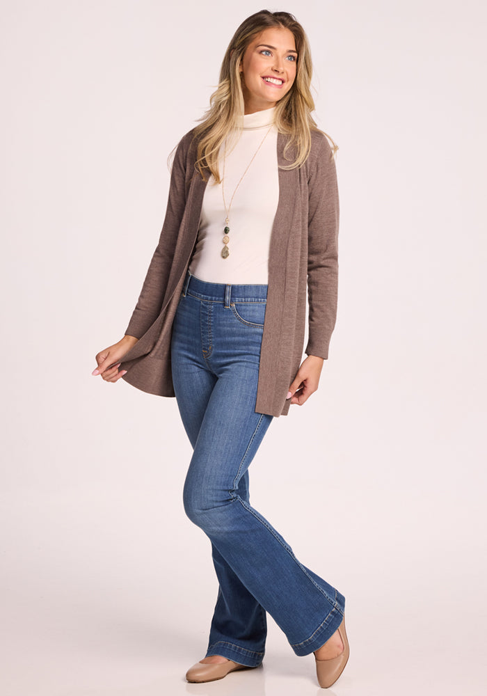 A smiling woman poses in the Woolx Macie Sweater in Simply Taupe, paired with blue flared jeans and beige shoes. She wears her hair long and wavy for a cozy look, set against a plain light background.