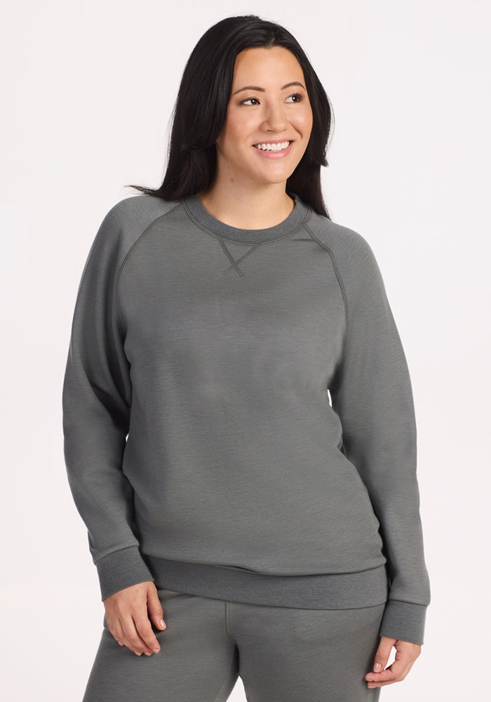 A woman with long dark hair smiles, wearing the Woolx Bailey Crewneck Sweatshirt in Sedona Sage with matching pants. She stands against a plain, light background, one hand by her side and the other on her hip. 