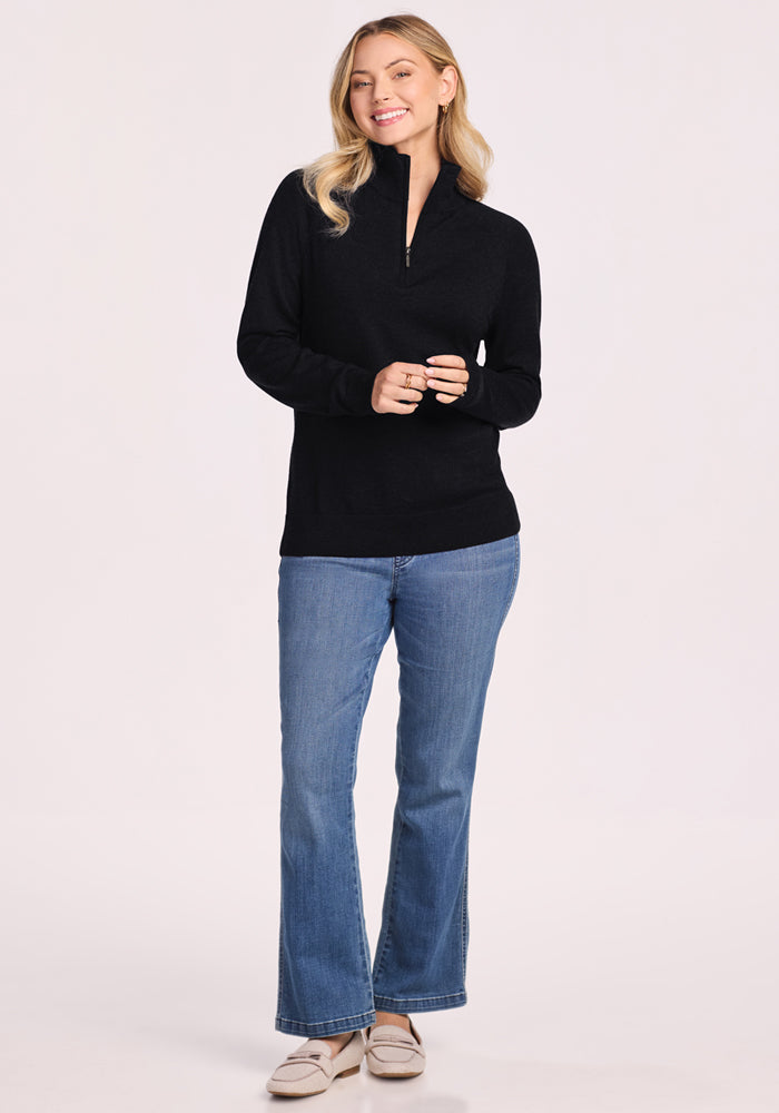 A woman with blonde hair smiles, wearing the Woolx Collins Half Zip Sweater in black, blue jeans, and light beige shoes. Her hands are loosely clasped in front of her against a plain, light-colored background.