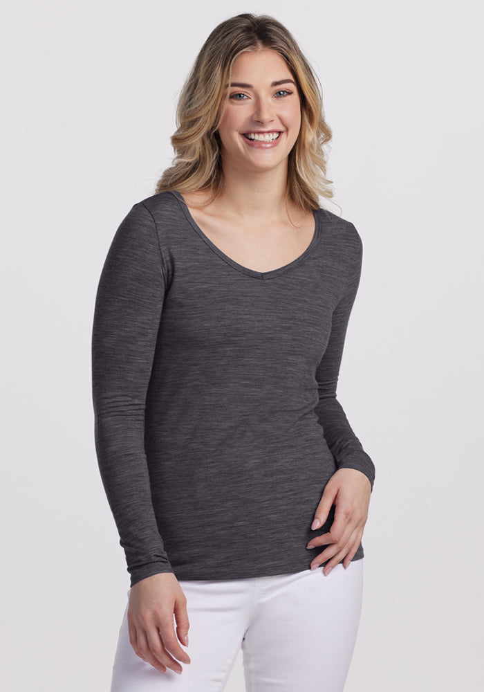 A woman with wavy blonde hair smiles while wearing the Woolx Layla V Neck Top in Cinder Melange, paired with white pants, standing against a plain white background. 