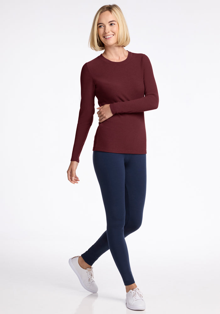 A smiling woman with short blonde hair wears the Woolx Hadley Ribbed Crew in Toasted Garnet, navy leggings, and white sneakers. Perfect for travel and lightweight layering, she stands against a plain white background with one leg crossed.
