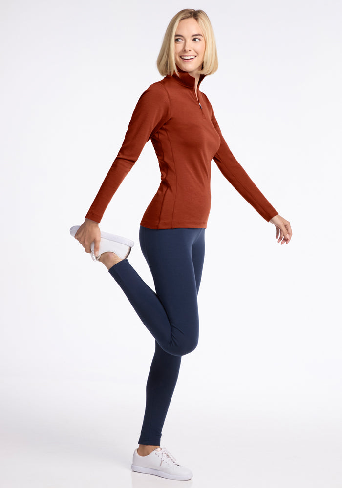 A woman wearing the Woolx Brooke 1/4 Zip in Maple Leaf and navy leggings stands on one leg, smiling as she stretches by holding her other foot behind her, looking slightly to the side.