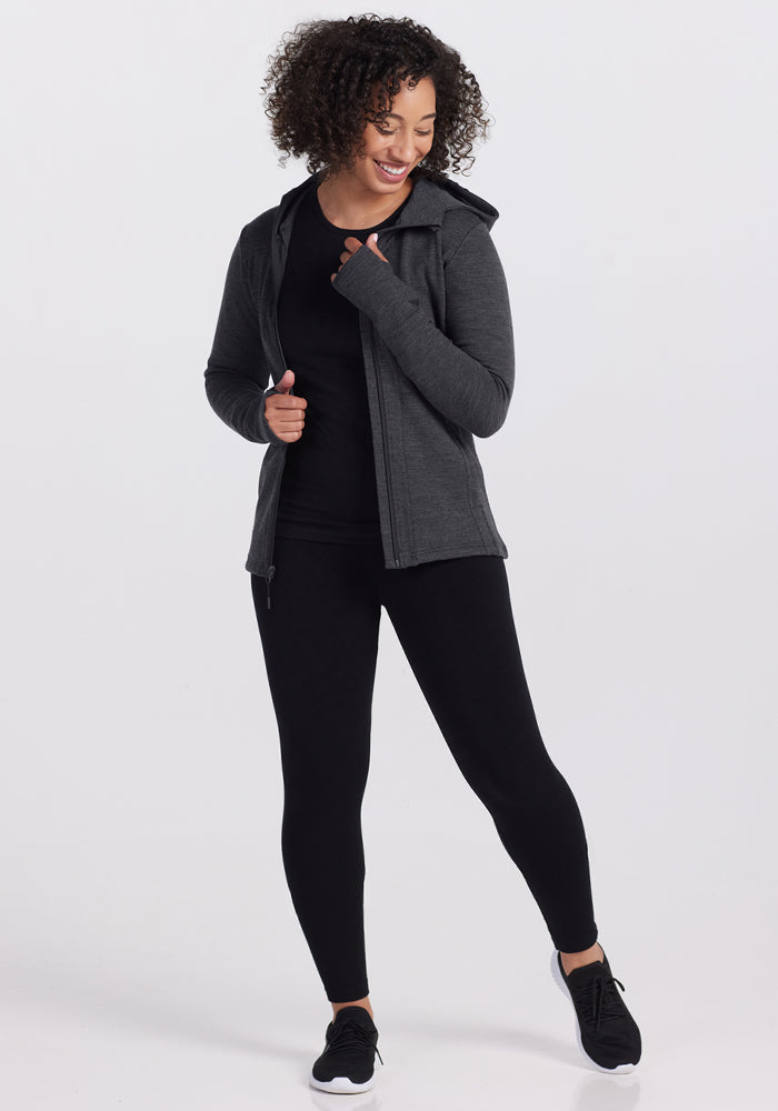 A woman with curly hair smiles while wearing a Woolx Zoey Zip Up Hoodie in Cinder Melange, black leggings, and black sneakers, standing against a plain white background.