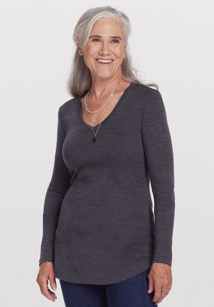 Smiling older woman with long gray hair, wearing the Woolx Eva Tunic in Cinder Melange, blue jeans, and layered necklaces, stands against a plain white background. 