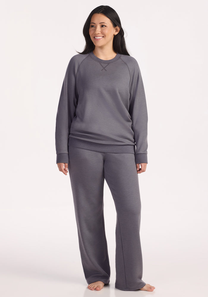 A woman with long dark hair smiles barefoot, wearing the Woolx Bailey Crewneck Sweatshirt in Arctic Dusk with matching gray pants, against a plain light background.