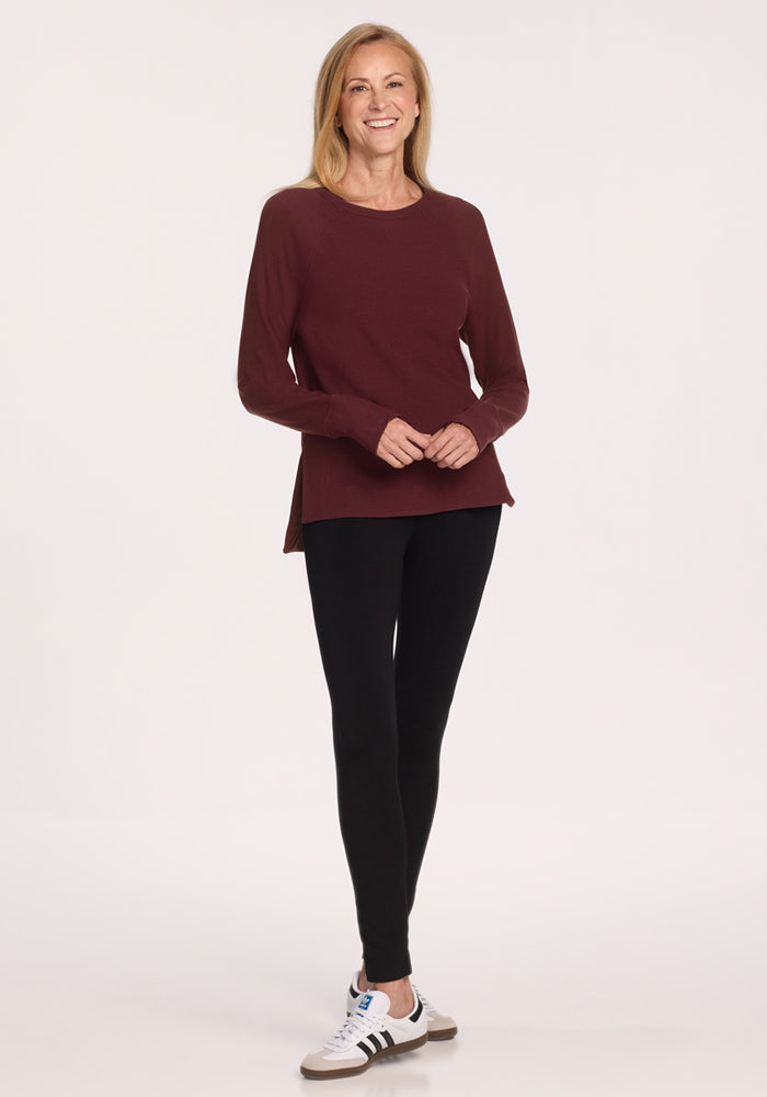 A smiling woman with long blonde hair wears the Woolx Ainsley Sweater in Toasted Garnet, paired with black leggings and white sneakers with black stripes, standing against a plain light background.