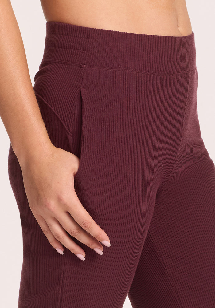 Someone wears the Woolx Luca Ribbed Pants in Toasted Garnet—high-waisted, wide-leg lounge pants with a soft vertical rib texture—resting their right hand on their hip against a plain, light-colored background.