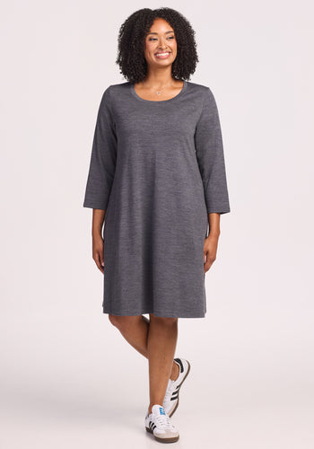 A smiling woman with curly hair wears the Woolx Rissa A Line Dress in Cinder Melange, styled with white sneakers against a light backdrop—ideal for fall/winter layering or as a cozy travel dress. | Maysa is 5’3”, wearing a size M
