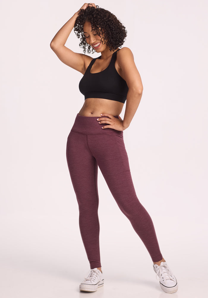A woman with curly hair smiles while posing in a black sports bra, Woolx Frost Duralite® Leggings in Grape Harvest Melange, and white sneakers against a plain light background.