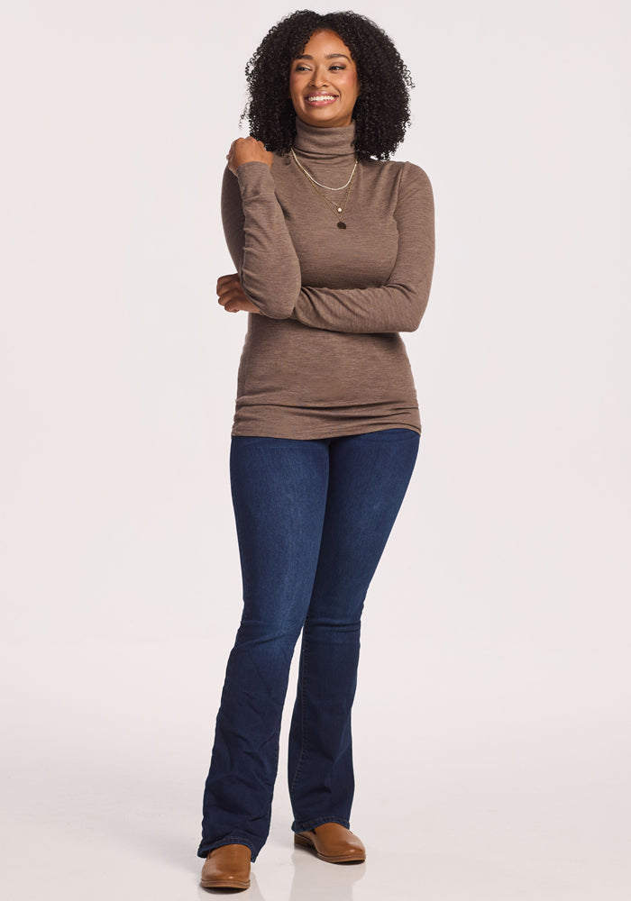 A woman smiles with arms crossed, wearing the Woolx Sage Ribbed Turtleneck in Simply Taupe, blue jeans, brown shoes, and layered necklaces. The background is plain white.