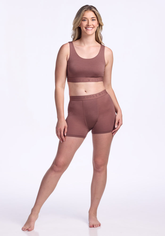 A woman stands barefoot against a white background, smiling, wearing the Woolx Aubrey Lounge Bra in Nutmeg with high-waisted shorts.