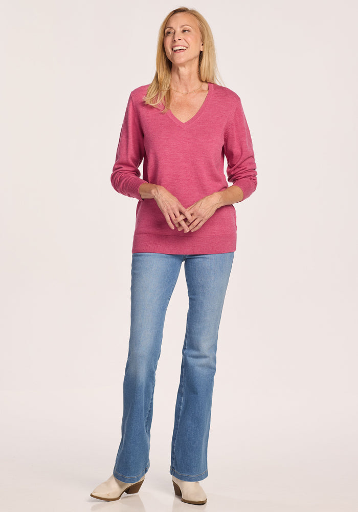 A smiling woman with long blonde hair wears the Woolx Francesca Sweater in Wild Salmon, paired with blue jeans and beige shoes, standing against a plain light background.
